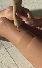 Close up of Golden Era Blurring Body Glow tube showcasing sandstone shimmer being applied to a tanned leg on a tile floor