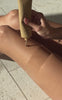 Close up of Golden Era Blurring Body Glow tube showcasing sandstone shimmer being applied to a tanned leg on a tile floor