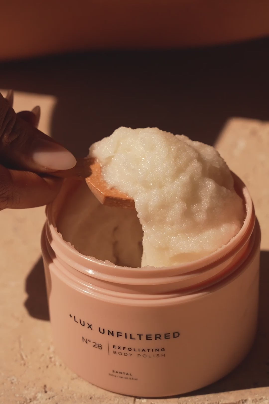 Hand scooping creamy exfoliating body polish from pink jar labeled +LUX UNFILTERED N°28 EXFOLIATING BODY POLISH SANTAL