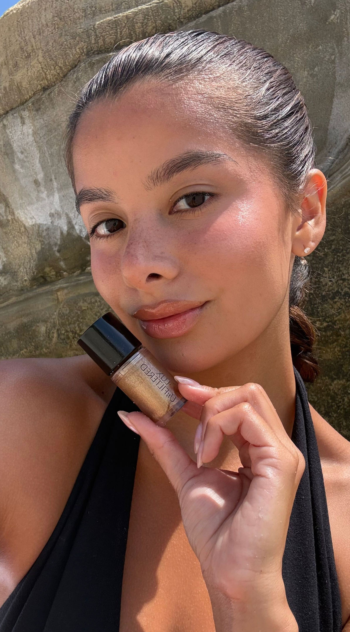 Woman holding a small bottle of shimmery sunless tanning drops near her cheek in sunlight with stone background