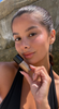 Woman holding a small bottle of shimmery sunless tanning drops near her cheek in sunlight with stone background