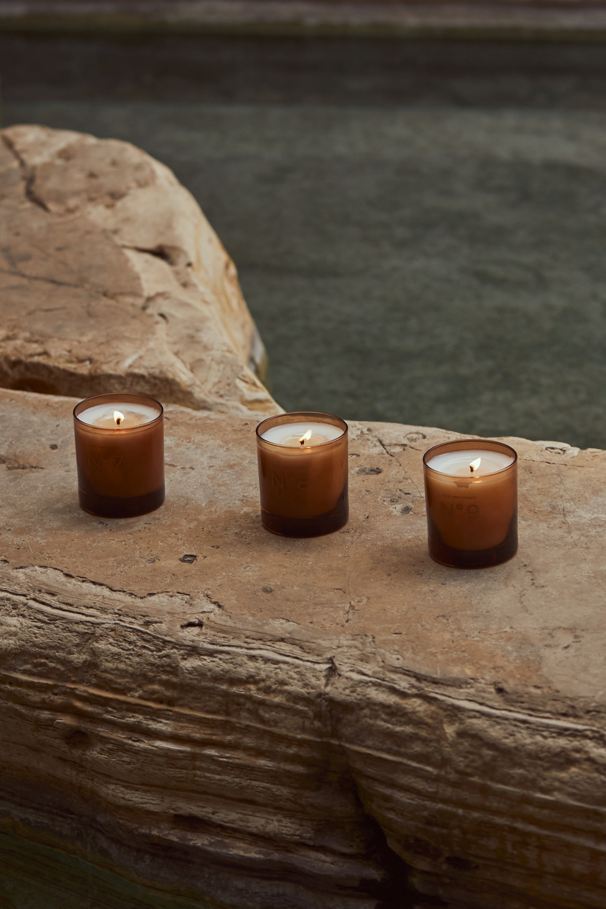 Three lit amber glass candles on a weathered stone ledge beside calm water